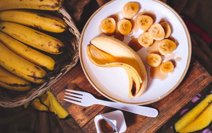 tavolo di legno con casto di legno con all'interno delle banane. Un piatto bianco con una banana intera ed una banana a fette. Un forchetta bianca insieme ad un vasetto di yogurt aperto